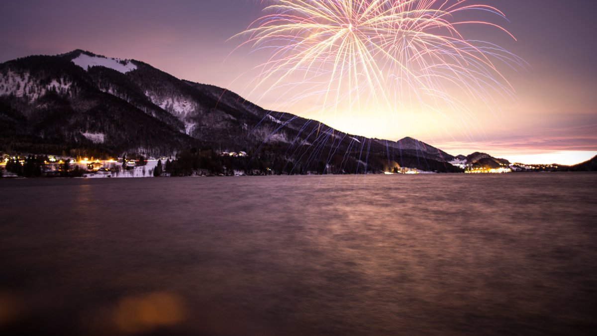 Farbenfrohes Feuerwerk am Nachthimmel über einem ruhigen See und umgeben von Bergen