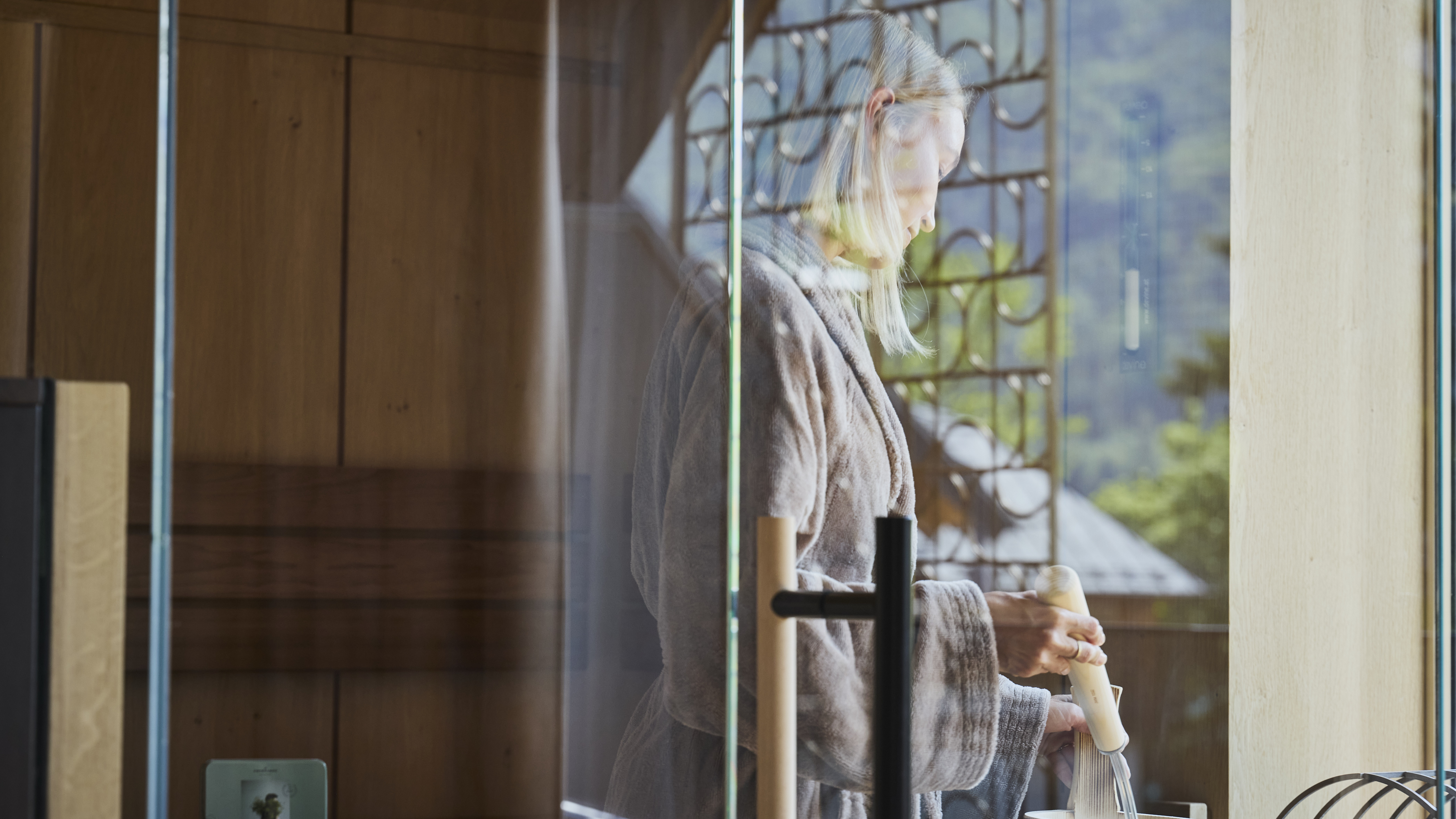 Individual in a robe inside a sauna reflecting in glass enclosure preparing for a treatment
