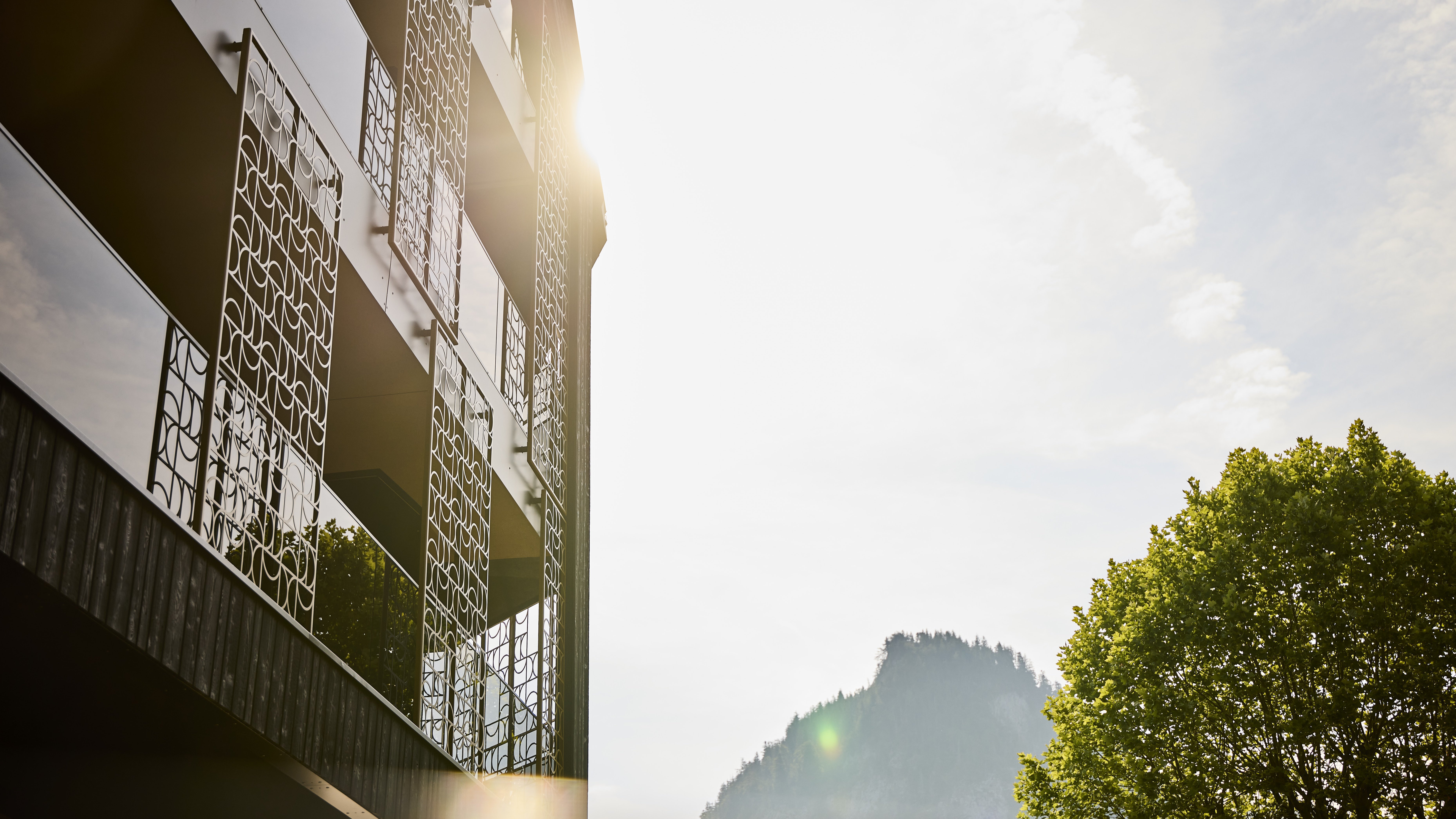 Modernes Gebäude mit Balkonen, beleuchtet von Sonnenlicht, umgeben von Bäumen und Bergen.